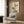 May include: Botanical wall art featuring pink poppies with green leaves and stems on cream background, displayed in light wood frame above beige upholstered armchair with geometric pillow. Modern reading nook with brass floor lamp, dark side table with open book, and warm wood paneling.