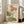 May include: Large framed botanical print leaning against white wall showing pink and white poppies with green leaves in vintage illustration style. Cream-colored artwork in light wood frame positioned next to beige upholstered chair near bright windows in modern living room setting.