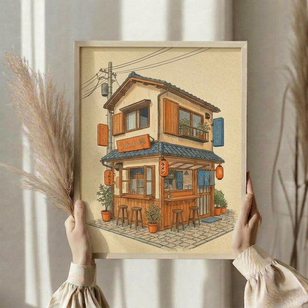 May include: Hands holding a framed illustration of a cozy two-story Japanese ramen shop with warm wood exterior, orange lanterns, outdoor seating with stools, potted plants, and Japanese text signage. The artwork has vintage beige tones and detailed architectural elements including traditional roof tiles and power lines. Dried wheat stalks visible to the left side.