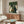May include: Framed poster showing six cats gathered around a round wooden table playing cards on green textured background. White, black, and tuxedo cats sit on wooden chairs holding cards with poker chips on table. Artwork hangs above mid-century walnut credenza in modern living room with cream upholstered chair, glass coffee table, and white table lamp.