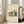 May include: A framed poster featuring a modernist architectural building illustration in muted green, orange and beige tones with "BAUHAUS" and "International Design Museum" text at bottom. The cream-colored print is displayed in a light wood frame, leaning against white walls near large windows in a bright living room with a beige sectional sofa and light hardwood floors.
