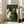 May include: Framed poster print displayed in bright living room showing six cats sitting around green card table playing poker. White and black cats hold playing cards with poker chips on table. Artwork features vintage woodblock print style with green textured background. Light wood frame leans against white wall near beige upholstered chair and large windows with natural sunlight streaming in.