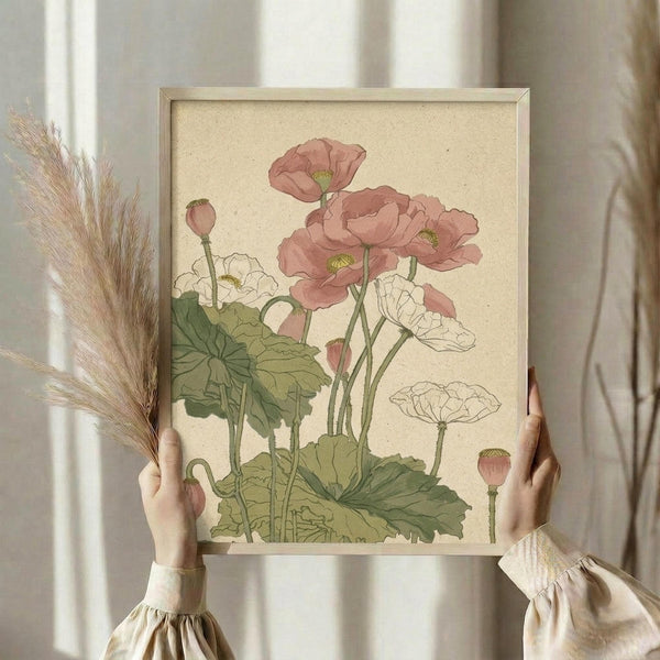 May include: Person in cream ruffled sleeves holding framed botanical illustration of pink and white poppies with green leaves against beige background. Dried pampas grass visible to left. Natural lighting from window creates soft shadows on neutral wall.
