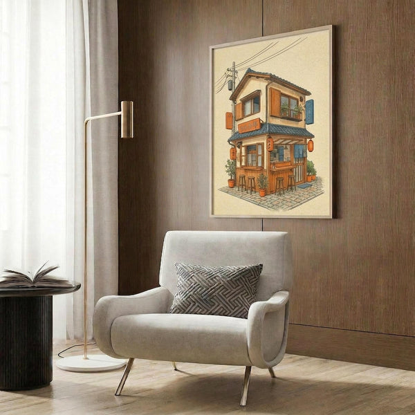 May include: Framed artwork showing a detailed illustration of a traditional Japanese two-story building with warm orange and blue wooden shutters, potted plants, and power lines, displayed on a wood-paneled wall above a cream upholstered armchair with geometric patterned pillow, beside a brass floor lamp and dark round side table with open book.