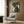 May include: Framed poster featuring cats playing cards around a green table in woodblock print style, displayed on wood paneled wall above beige upholstered chair with geometric pillow. Room shows modern brass floor lamp, dark side table with open book, and neutral decor with natural lighting from window.