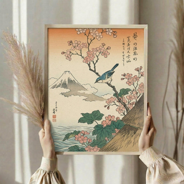 May include: Person in cream clothing holding framed Japanese woodblock print featuring blue bird perched on cherry blossom branch with Mount Fuji in background, surrounded by pink flowers and green leaves, displayed against white curtained window with dried pampas grass visible to left side.