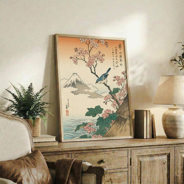 May include: Vintage Japanese woodblock print featuring Mount Fuji in background with cherry blossom tree, blue bird perched on branch, and flowing water below. Print displayed in light wooden frame on rustic wood sideboard alongside woven basket with fern plant, ceramic vase, and books. Warm neutral room setting with cream chair visible.