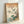 Framed Japanese woodblock print artwork leaning against white wall, featuring Mount Fuji in background with clouds, cherry blossom branches with pink flowers, blue bird perched on branch, green leaves, flowing water at bottom, and Japanese calligraphy text on cream-colored paper in light wooden frame