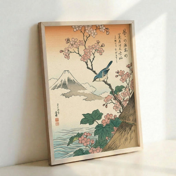 Framed Japanese woodblock print artwork leaning against white wall, featuring Mount Fuji in background with clouds, cherry blossom branches with pink flowers, blue bird perched on branch, green leaves, flowing water at bottom, and Japanese calligraphy text on cream-colored paper in light wooden frame