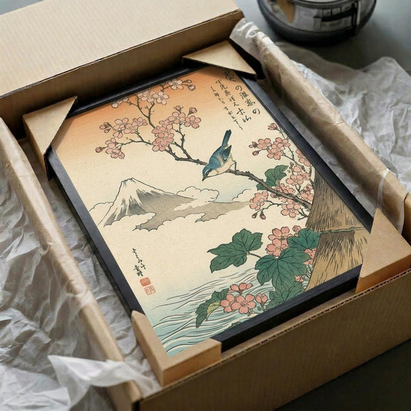 May include: Japanese-style woodblock print artwork displayed in black frame inside cardboard shipping box with protective wrapping. The framed print shows a small blue bird perched on cherry blossom branch with pink flowers, Mount Fuji visible in background through clouds, green leaves and flowing water below. Traditional ukiyo-e art style with muted earth tones and Japanese calligraphy text.