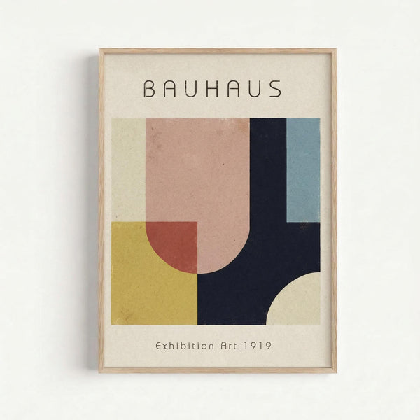 May include: Framed poster with light wood frame hanging on white wall. Features geometric abstract design with overlapping shapes in muted colors including yellow, coral pink, sage green, navy blue, and cream. Text reads "BAUHAUS" at top and "Exhibition Art 1919" at bottom on cream background.