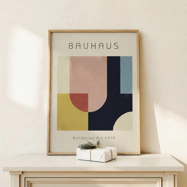 May include: Framed Bauhaus-style art print with geometric shapes in muted colors including pink, yellow, navy blue, and sage green, displayed on light wood surface next to small wrapped gift with twine and evergreen sprig against cream wall