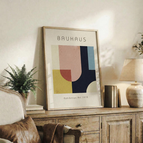 May include: Framed Bauhaus-style geometric art print in light wood frame, displayed on rustic wooden sideboard alongside fern plant, books, and ceramic vase. Abstract composition features overlapping shapes in pink, yellow, navy blue, sage green, and cream against white background. Cozy living room setting with leather armchair and neutral textiles.