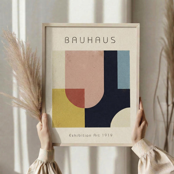 May include: Person holding framed Bauhaus-style geometric art print with abstract shapes in mustard yellow, coral pink, navy blue, and light blue colors. Dried pampas grass visible on left side. Light wooden frame against neutral beige background with soft natural lighting.