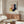 May include: Framed Bauhaus-style poster with geometric shapes in pink, yellow, navy, and blue hanging above a wooden credenza. Modern living room setting with white curved chair, round glass coffee table, mushroom table lamp, and floor lamp. Warm natural lighting creates shadows on cream walls.