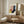 May include: Framed Bauhaus-style poster with geometric shapes in pink, yellow, coral, navy blue and teal displayed on wooden sideboard in modern living room. White upholstered chair, brass table lamp with cream shade, and small round wooden side table complete the minimalist Scandinavian-inspired interior setting.