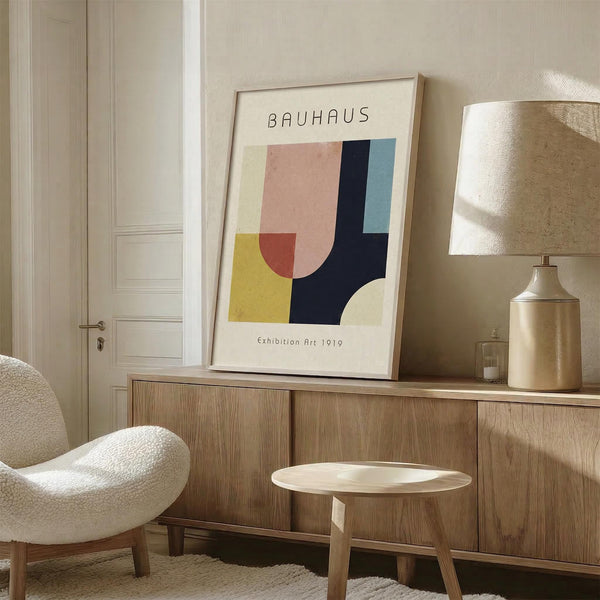 May include: Framed Bauhaus-style poster with geometric shapes in pink, yellow, coral, navy blue and teal displayed on wooden sideboard in modern living room. White upholstered chair, brass table lamp with cream shade, and small round wooden side table complete the minimalist Scandinavian-inspired interior setting.