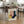 May include: Framed Bauhaus-style geometric poster with abstract shapes in pink, yellow, navy blue, and cream colors, leaning against wooden dining table leg in modern kitchen. Natural lighting illuminates the minimalist artwork among woven chairs, spider plant, and white cabinetry on hardwood floors.