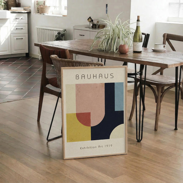 May include: Framed Bauhaus-style geometric poster with abstract shapes in pink, yellow, navy blue, and cream colors, leaning against wooden dining table leg in modern kitchen. Natural lighting illuminates the minimalist artwork among woven chairs, spider plant, and white cabinetry on hardwood floors.