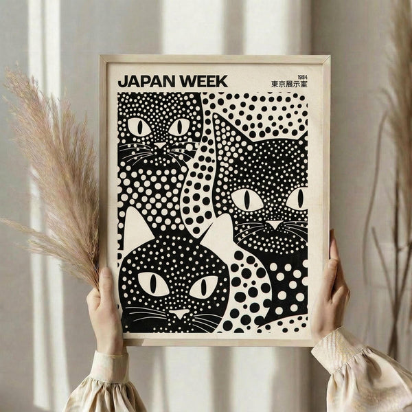 May include: Person in cream sweater holding framed black and white polka dot poster featuring stylized cat faces with whiskers and geometric patterns. Light wood frame against neutral curtained background with dried wheat stems visible on left side.