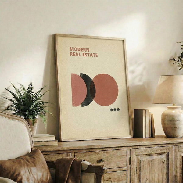 May include: Framed poster with cream background displaying brown and black geometric circles above three small dots, leaning against white wall on rustic wooden sideboard beside fern plant, books, ceramic vases, and partial view of beige upholstered chair