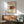 May include: Modern living room with sage green upholstered chair, wooden sideboard, and white table lamp. Framed poster on wall features abstract overlapping circles in coral and dark green on cream background with "MODERN REAL ESTATE" text. Natural sunlight creates dramatic shadows across white wall and furniture.