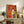 May include: Framed colorful abstract dog illustration artwork displayed on rustic wooden sideboard in modern living room setting. Four stylized dog silhouettes in vibrant orange, blue, yellow, and red geometric shapes with textured brushstroke background. Light wood frame, positioned beside fern plant, books, and ceramic pottery on weathered console table with beige armchair visible.