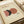 Modern minimalist poster with abstract geometric design featuring overlapping shapes in terracotta, dusty rose, and dark charcoal colors on cream background. Text reads "MODERN REAL ESTATE" in rust-colored letters. Print is framed in light wood frame and photographed at an angle on neutral surface with partial roll of brown paper visible.
