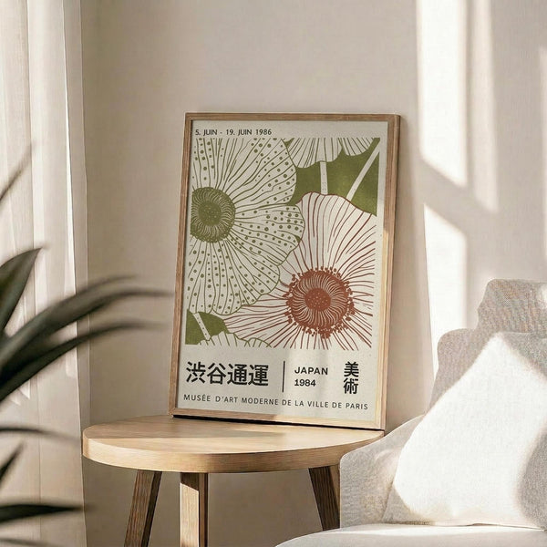 May include: Framed botanical poster with stylized sunflowers in green and terracotta colors, featuring Japanese text and museum exhibition details from 1984. Poster sits on round wooden side table next to beige upholstered chair with white throw pillow. Plant leaves visible in corner, soft natural lighting creates shadows on neutral wall background.