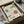 May include: Framed botanical poster featuring stylized flowers in green and coral colors with Japanese text, displayed in black frame partially unwrapped from brown cardboard packaging with tissue paper, surrounded by wooden dowels on white fabric surface.