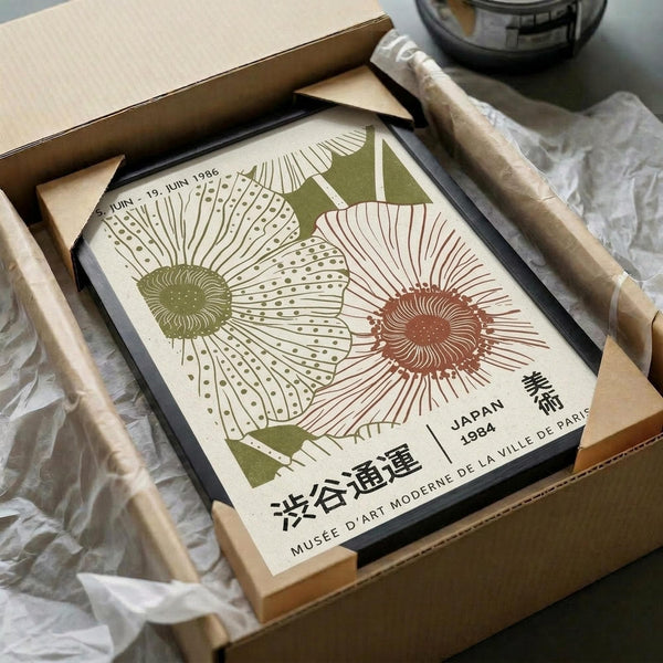 May include: Framed botanical poster featuring stylized flowers in green and coral colors with Japanese text, displayed in black frame partially unwrapped from brown cardboard packaging with tissue paper, surrounded by wooden dowels on white fabric surface.