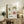 May include: Framed botanical poster with stylized sunflowers in green and coral colors displayed on rustic wooden sideboard in cozy living room. Features Japanese and French text, dates from 1986. Scene includes fern plant, ceramic vases, stacked books, and beige upholstered furniture with throw pillow.