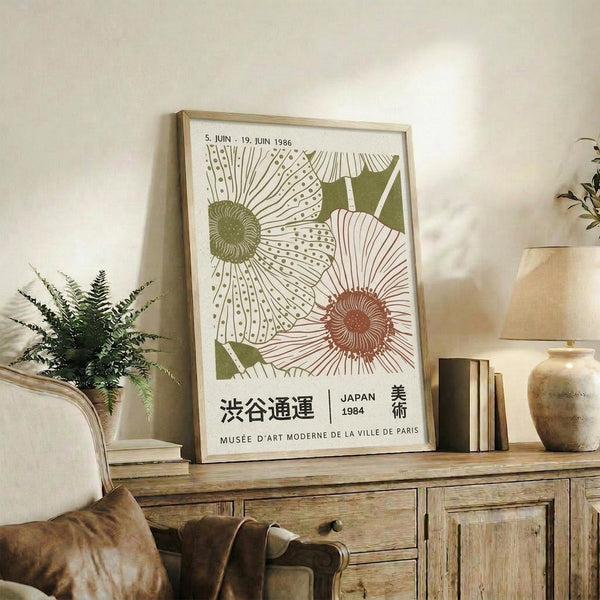 May include: Framed botanical poster with stylized sunflowers in green and coral colors displayed on rustic wooden sideboard in cozy living room. Features Japanese and French text, dates from 1986. Scene includes fern plant, ceramic vases, stacked books, and beige upholstered furniture with throw pillow.