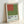 May include: Framed exhibition poster featuring a minimalist geometric design with an orange-red square positioned on a larger olive green square against a white background. The poster displays "Ellsworth Kelly" and "The Detroit Institute of Arts" text with exhibition dates "June 25 - August 11" at the bottom. Light wood frame leaning against a white wall, showing canvas texture throughout the print.