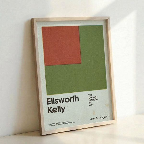 May include: Framed exhibition poster featuring a minimalist geometric design with an orange-red square positioned on a larger olive green square against a white background. The poster displays "Ellsworth Kelly" and "The Detroit Institute of Arts" text with exhibition dates "June 25 - August 11" at the bottom. Light wood frame leaning against a white wall, showing canvas texture throughout the print.