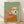 May include: Framed artwork leaning against white wall on light wooden floor. The painting shows a white fluffy terrier dog wearing yellow goggles, coral pink hood and flowing scarf against sage green background. Artwork has natural wood frame and painterly brushstroke texture. Soft natural lighting creates gentle shadows on the white textured wall behind.