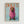 A dachshund wearing a pink knitted hoodie sits against a light blue background on a framed poster print. The dog's brown and tan face peeks out from the ribbed pink sweater that covers its head and body. Orange text reading "DOGUE" appears at the top in a magazine-style layout. The artwork is displayed in a light wood frame against a white wall.