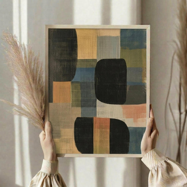 May include: Person in beige ruffled sleeves holding framed abstract geometric artwork featuring overlapping rectangular and rounded shapes in muted tones of sage green, dusty blue, coral pink, cream, and black with visible brushstroke texture. Dried pampas grass visible to left against soft natural lighting and neutral curtained background.