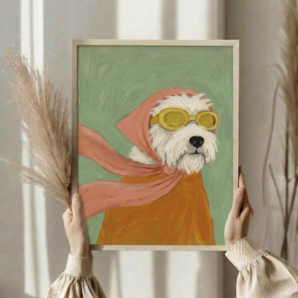 May include: Person holding framed artwork featuring a white terrier dog wearing yellow goggles and a pink flowing scarf against a sage green background, painted in an illustrative style. The painting is displayed in a light wood frame, with dried pampas grass visible to the left and soft natural lighting from nearby windows.