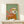 May include: Framed painting of a white terrier dog wearing yellow goggles and a flowing pink scarf against a sage green background, displayed on a light wood console table next to a small wrapped gift with evergreen sprig, against a cream wall