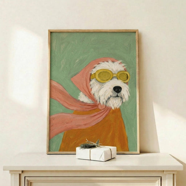 May include: Framed painting of a white terrier dog wearing yellow goggles and a flowing pink scarf against a sage green background, displayed on a light wood console table next to a small wrapped gift with evergreen sprig, against a cream wall
