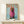 May include: Framed poster print featuring a brown dachshund wearing a pink knit hoodie on a light blue background, styled as a fashion magazine cover with "DOGUE" in orange letters. The wooden-framed artwork is displayed on a light wood shelf beside a small white gift box tied with twine and greenery, against a white wall.