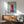 May include: Framed poster print featuring a dachshund dog wearing a bright pink knitted hooded sweater against a turquoise background, with "DOGUE" text in orange letters at top. Print displayed above mid-century modern wood credenza with white ceramic lamp, next to sage green upholstered armchair with wooden frame in bright, sunlit room with white walls.