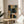 May include: Abstract geometric artwork in wooden frame displayed on round wooden side table next to beige upholstered chair. The framed print features overlapping rectangular shapes in muted tones of sage green, dusty blue, coral orange, cream, and charcoal black with visible canvas texture. Natural lighting creates soft shadows on white wall, with plant leaves visible in corner of serene modern living room setting.
