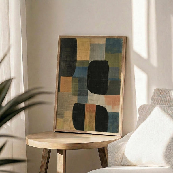 May include: Abstract geometric artwork in wooden frame displayed on round wooden side table next to beige upholstered chair. The framed print features overlapping rectangular shapes in muted tones of sage green, dusty blue, coral orange, cream, and charcoal black with visible canvas texture. Natural lighting creates soft shadows on white wall, with plant leaves visible in corner of serene modern living room setting.