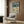 May include: Vintage-style travel poster featuring colorful birds perched on cherry blossom branches against pale green background, with "JAPAN 1973" and Japanese characters at top. Displayed in light wood frame on textured brown wall above beige upholstered armchair with geometric patterned pillow. Modern brass floor lamp and side table with open book complete the contemporary reading nook scene.