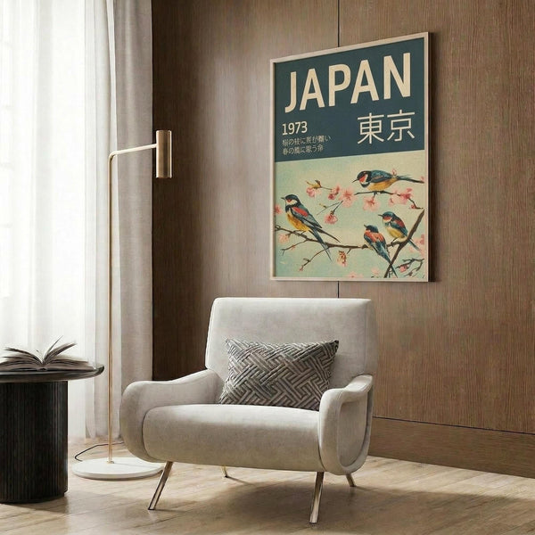 May include: Vintage-style travel poster featuring colorful birds perched on cherry blossom branches against pale green background, with "JAPAN 1973" and Japanese characters at top. Displayed in light wood frame on textured brown wall above beige upholstered armchair with geometric patterned pillow. Modern brass floor lamp and side table with open book complete the contemporary reading nook scene.