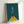 May include: Framed minimalist bicycle poster with teal background and yellow triangular base, showing white and black geometric bicycle handlebars and stem in simple line art style, displayed in light wood frame leaning against white wall