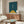 May include: Modern living room with cream walls showcasing a framed abstract geometric poster featuring teal blue background, yellow triangle, and white vertical bicycle form with black linear details. Styled with walnut wood credenza, white sculptural chair, oval glass coffee table, and contemporary lighting fixtures.