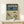 May include: Vintage-style travel poster featuring Japan with teal green header and cream text reading "JAPAN 1973" and Tokyo in Japanese characters. Four colorful birds with blue, orange and yellow plumage perch on cherry blossom branches with delicate pink flowers against a pale mint background. Displayed in light wood frame on white wooden shelf next to small white wrapped gift with evergreen sprig.